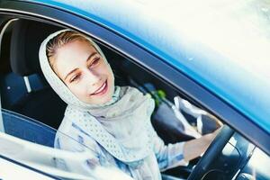 hermosa joven musulmán mujer en hijab conducción un azul coche solo y sonriente foto