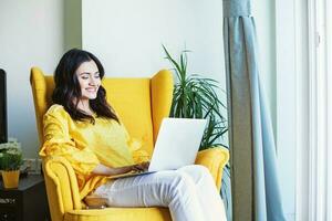 Young Indian woman using her laptop at home while sitting on a yellow armchair photo