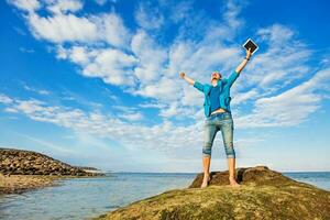 a young caucasian man standing on a rock by beach with his arms open looking in sky photo