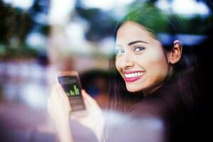 A beautiful young Indian woman smiling and looking through a glass window while holding a phone with graphs in her hands photo