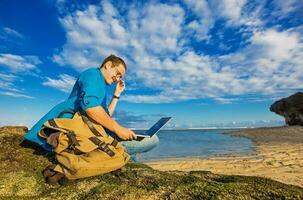 young caucasian man sitting on a beach working on a laptop with a bag lying by his side photo