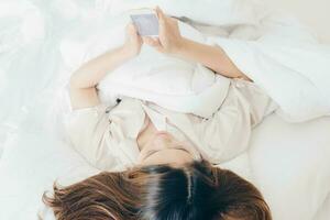 asiático mujer acostado en cama y relajante por utilizando su inteligente teléfono. foto