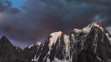 Darkness mountain landscape with great snowy mountain lit by dawn sun among dark clouds. Awesome alpine scenery with high mountain pinnacle at sunset or at sunrise. Big glacier on top in orange light. photo