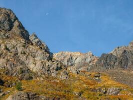 Bright mountain landscape with sharp rocks above yellow slope under blue sky with moon in sunny day. Colorful scenery with gold sunlit sharp rocky mountains. High rocky mountains in golden sunlight. photo