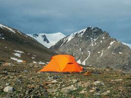 Orange tent on a sunny mountain slope. Camping on a rocky high-altitude plateau. Tent on the background of glacier and high snow-capped mountains. Solo trekking. photo