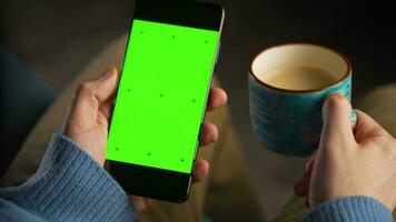 man använder sig av smartphone med grön mock-up skärm i vertikal läge och dricka kaffe. man bläddring internet, tittar på innehåll, videoklipp video