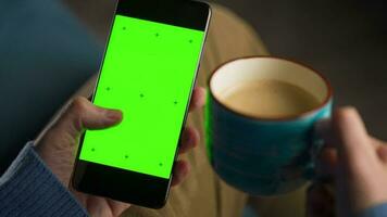 Mens gebruik makend van smartphone met groen mock-up scherm in verticaal mode en drinken koffie. Mens browsen internetten, aan het kijken inhoud, videos