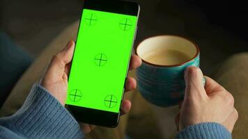 uomo utilizzando smartphone con verde modello schermo nel verticale modalità e potabile caffè. uomo navigazione Internet, Guardando contenuto, video