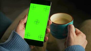 homme en utilisant téléphone intelligent avec vert maquette écran dans verticale mode et en buvant café. homme navigation l'Internet, en train de regarder contenu, vidéos video