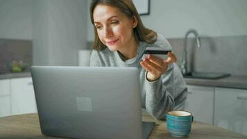 mujer en pie en hogar acogedor cocina, propensión terminado el mesa y hace un en línea compras utilizando un crédito tarjeta y ordenador portátil video