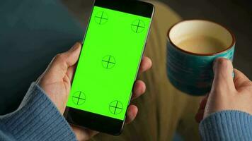 man använder sig av smartphone med grön mock-up skärm i vertikal läge och dricka kaffe. man bläddring internet, tittar på innehåll, videoklipp video