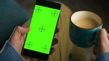 man använder sig av smartphone med grön mock-up skärm i vertikal läge och dricka kaffe. man bläddring internet, tittar på innehåll, videoklipp video