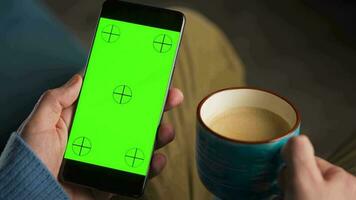 Mens gebruik makend van smartphone met groen mock-up scherm in verticaal mode en drinken koffie. Mens browsen internetten, aan het kijken inhoud, videos