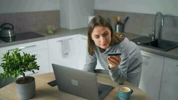 mujer en pie en hogar acogedor cocina, propensión terminado el mesa y hace un en línea compras utilizando un crédito tarjeta y ordenador portátil video