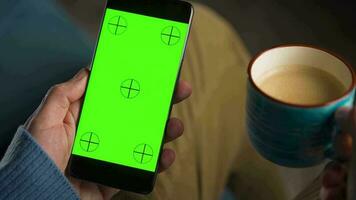 Mens gebruik makend van smartphone met groen mock-up scherm in verticaal mode en drinken koffie. Mens browsen internetten, aan het kijken inhoud, videos