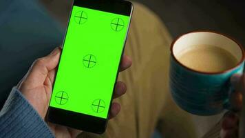 Mann mit Smartphone mit Grün Attrappe, Lehrmodell, Simulation Bildschirm im Vertikale Modus und Trinken Kaffee. Mann Surfen Internet, Aufpassen Inhalt, Videos