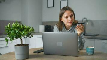 vrouw staand in huis knus keuken, leunend over- de tafel en maakt een online boodschappen doen gebruik makend van een credit kaart en laptop video