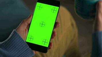 man använder sig av smartphone med grön mock-up skärm i vertikal läge och dricka kaffe. man bläddring internet, tittar på innehåll, videoklipp video