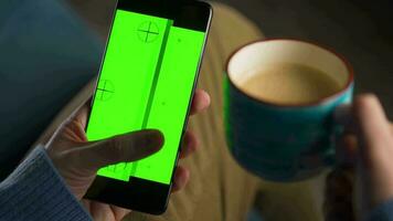 man använder sig av smartphone med grön mock-up skärm i vertikal läge och dricka kaffe. man bläddring internet, tittar på innehåll, videoklipp video