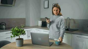 mujer con taza de café en mano en pie en hogar acogedor cocina utilizar ordenador portátil hacer Lanza libre trabajar, chateando con alguien, respondiendo correo electrónico video