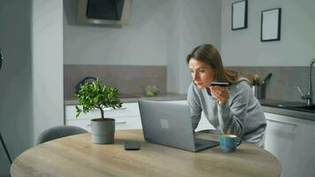 vrouw staand in huis knus keuken, leunend over- de tafel en maakt een online boodschappen doen gebruik makend van een credit kaart en laptop video