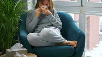 Unhealthy Caucasian woman sits in a chair and sneezes or blows her nose into a napkin because she has a cold, flu, coronavirus. It is snowing outside video