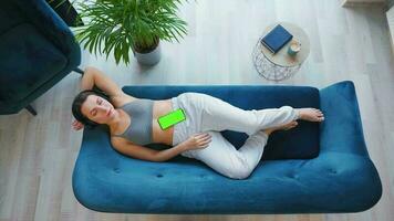 Overhead shot of relaxed woman in wireless headphones listening to music and using mobile apps on smartphone with green mock-up screen video