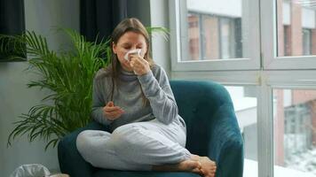 Unhealthy Caucasian woman sits in a chair and sneezes or blows her nose into a napkin because she has a cold, flu, coronavirus. It is snowing outside video