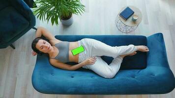 Overhead shot of relaxed woman in wireless headphones listening to music and using mobile apps on smartphone with green mock-up screen video