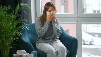 Unhealthy Caucasian woman sits in a chair and sneezes or blows her nose into a napkin because she has a cold, flu, coronavirus. It is snowing outside video