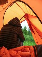 Loneliness in the tent. Man sits on the back in hiking dress in orange tent on mountain. Loneliness man tourist backpacker relax and enjoy beautiful nature outdoor lifestyle and holiday vacation. photo