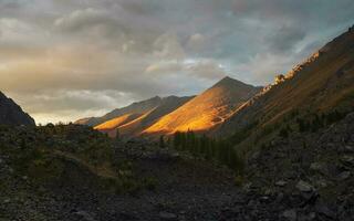 Panoramic colorful mountain landscape with great mountain lit by dawn sun among dark clouds. Awesome alpine scenery with high mountain pinnacle at sunset or at sunrise. Big mountain in orange light photo