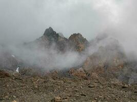 Atmospheric ghostly landscape with fuzzy silhouettes of sharp rocks in low clouds. Dramatic view to large mountains blurred in rain haze in gray low clouds. photo