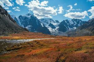 Wonderful alpine landscape with mountain river in valley in autumn colors on background of snowy mountains silhouettes under blue cloudy sky. Beautiful mountain valley in autumn. photo