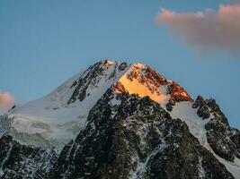 Top black rocks in evening golden sunshine and white-snow pointy peak. Minimalistic atmospheric landscape with high snowy mountain with peaked top under cirrus clouds in sky. photo