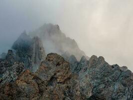 Atmospheric ghostly landscape with fuzzy silhouettes of sharp rocks in low clouds. Dramatic view to large mountains blurred in rain haze in gray low clouds. photo