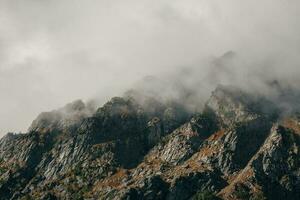 Dramatic fog among giant rocky mountains. Ghostly atmospheric view to big cliff in cloudy sky. Low clouds and beautiful rockies. Minimalist scenery mysterious place. photo