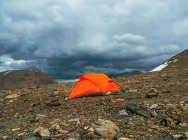 verano cámping en montaña. brillante alpino paisaje con vívido naranja tienda a muy alto altitud con ver a alto montaña y grande glaciar en dramático nubes increíble montaña paisaje con carpa. foto