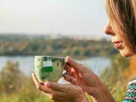 Women hands hold a porcelain mug with a bag inside, drink hot green tea at nature. Woman enjoy warm brew or beverage in cup, relax rest having break outside photo
