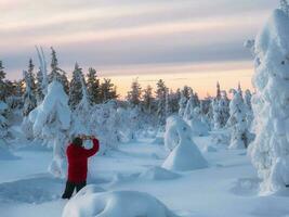 Man stands with his back in deep snow and taking photo using a smartphone the stunning views of snow-covered trees during a cold polar dawn. Winter vacation concept, trip to the Arctic fairy forest.