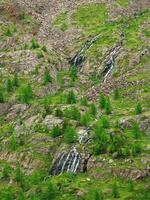 cascada mangas escénico distancia alpino paisaje con arboles en Pendiente verde rocoso grande montaña rango con cascada en soleado ligero. vistoso natural paisaje con un cascada en un escarpado acantilado. foto