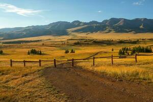 Dirt road is blocked by a fence. The path is closed, private territory. Beautiful sunny landscape with golden vast field with long fence. Vivid scenery with autumn field behind wood fence in mountains photo