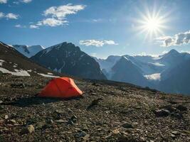 Tent on the edge of a cliff. Camping on a rocky high-altitude plateau. Orange tent on the background of high snow-capped mountains. photo