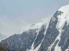 Winter atmospheric mountains landscape with blizzard in big snowy mountain top in low clouds. Awesome minimal scenery with white glacier on black rocks. High mountain pinnacle with snow in clouds. photo