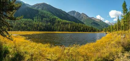 Panoramic view of sunny swampy autumn plateau. Awesome highland scenery with beautiful glacial lake among sunlit hills and rocks against mountain range. photo