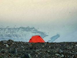 Sunlight slides across the surface of the glacier early in the morning. Camping high in the mountains. An orange tent on a rocky plateau on the background of a glacier. photo