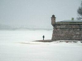 A lonely man walking, behind. Beautiful spring cityscape with snowfall. Contrasting view of the Peter and Paul Fortress in winter. Saint-Petersburg. photo