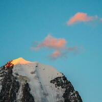 Top black rocks in evening golden sunshine and white-snow pointy peak. Atmospheric landscape with high snowy mountain with peaked top under cirrus clouds in sky. Square view. photo
