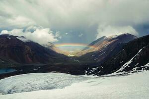 Rainbow over a mountain valley. Gloomy scenery with bright rainbow above glacier in mountain valley. Top view to colorful rainbow and low clouds in mountains in rainy and sunny weather. photo