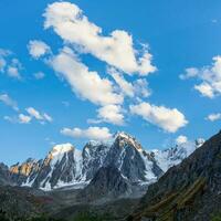 White clouds over a high pointed mountain. Snowy high-altitude alpine landscape with snow-capped mountain peak and sharp rocks under cloudy blue sky. photo